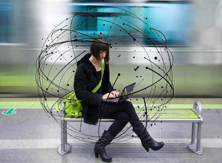 A young woman sitting on a bench at a train platform, working on a laptop.