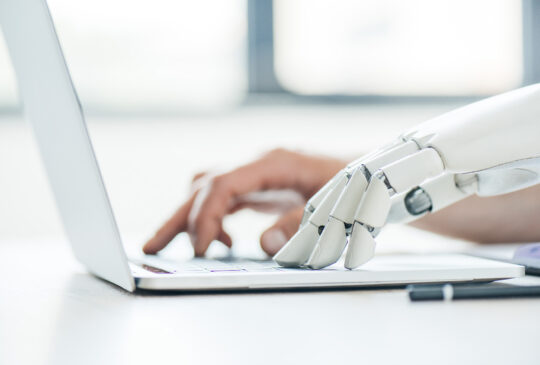 Menschliche Hand und Roboterhand tippen gemeinsam auf einer Laptop-Tastatur – Symbolbild für die Zusammenarbeit von Künstlicher Intelligenz und menschlichem Post-Editing.