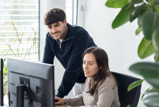 Zwei Mitarbeitende arbeiten gemeinsam an einem Computer im Büro und besprechen Inhalte am Bildschirm.