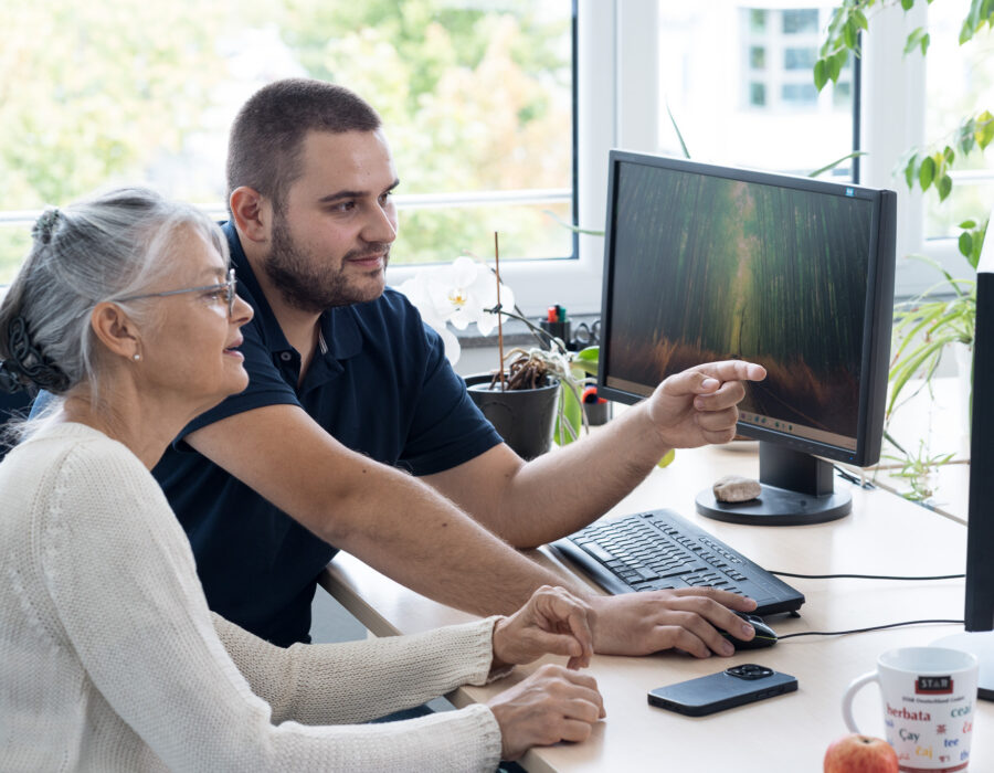Zwei Personen sitzen an einem Schreibtisch und betrachten gemeinsam Inhalte auf einem Computerbildschirm.