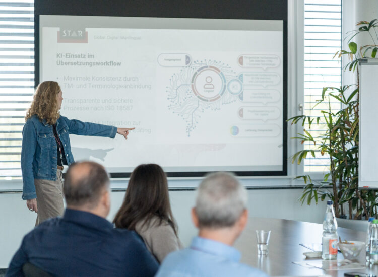 Person steht in einem Besprechungsraum vor einer Projektionsfläche und zeigt auf eine Folie, während mehrere Teilnehmende am Tisch zuhören.