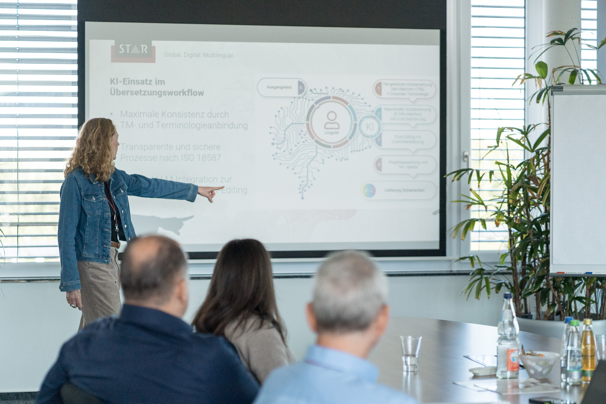 Person standing in a meeting room pointing at a projected slide while several attendees seated at a table listen.