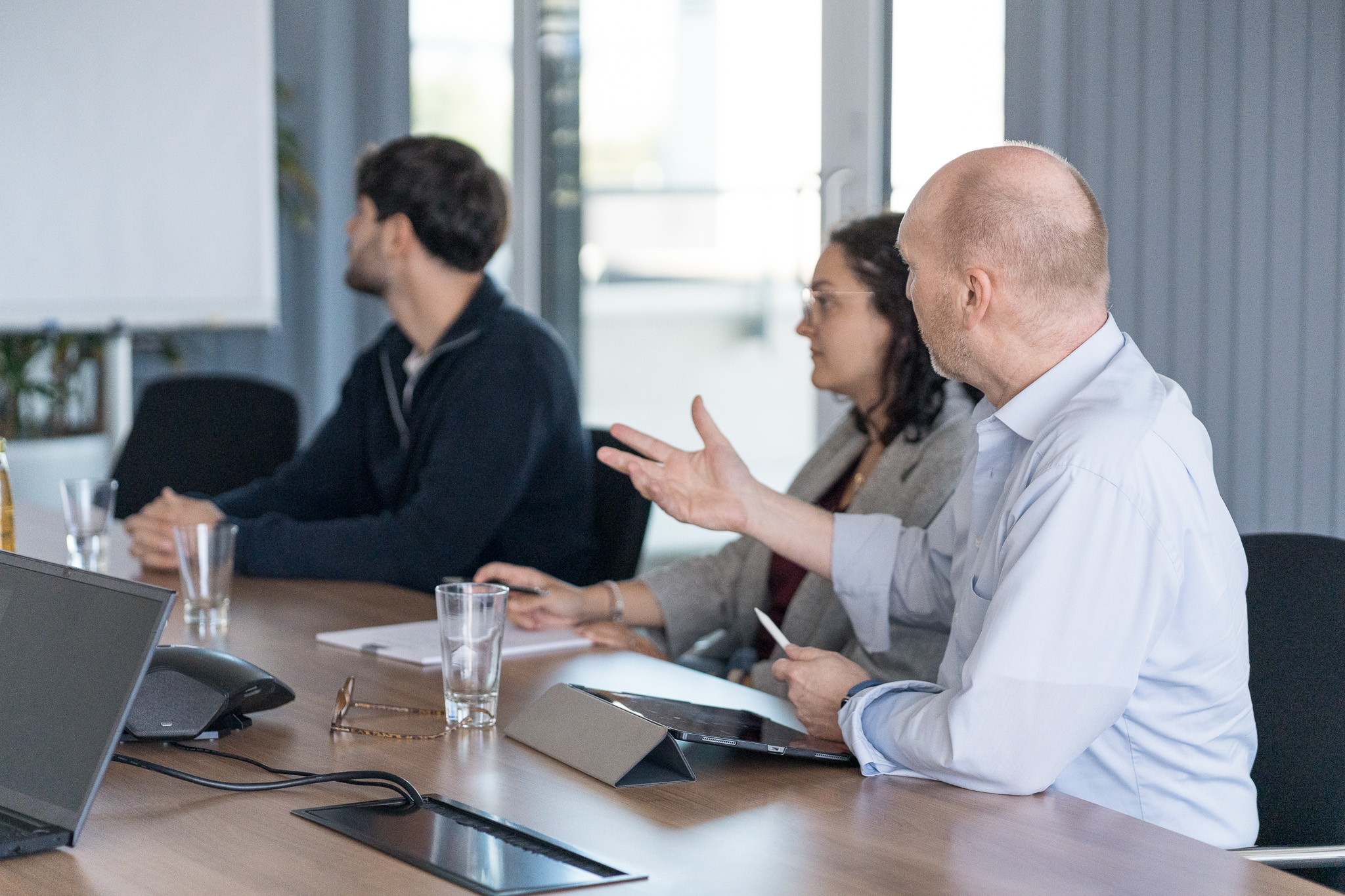 Mehrere Personen sitzen an einem Konferenztisch und sprechen während einer Besprechung, während im Hintergrund eine Präsentation zu sehen ist.