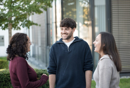 Drei Personen stehen draußen vor einem Gebäude und unterhalten sich miteinander.