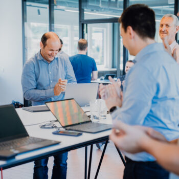 Several people gathered around a table with laptops applauding while one person works on a laptop, representing a successful project outcome in a modern work environment.